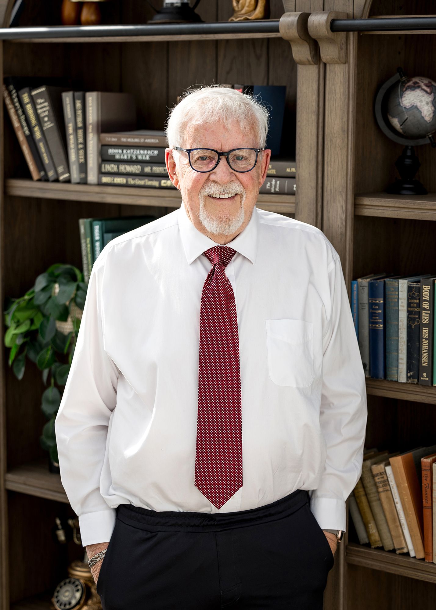 Smiling senior man with white hair and glasses wearing a white shirt and red tie, standing in front of a bookshelf.