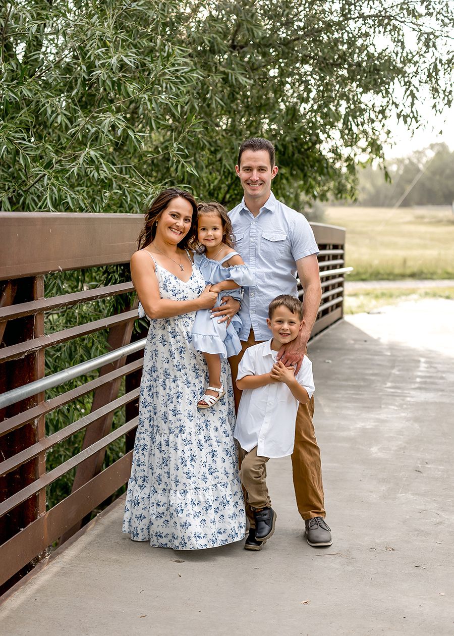 Happy family of four posing on a wooden bridge outdoors, parents with two young children in summer attire.