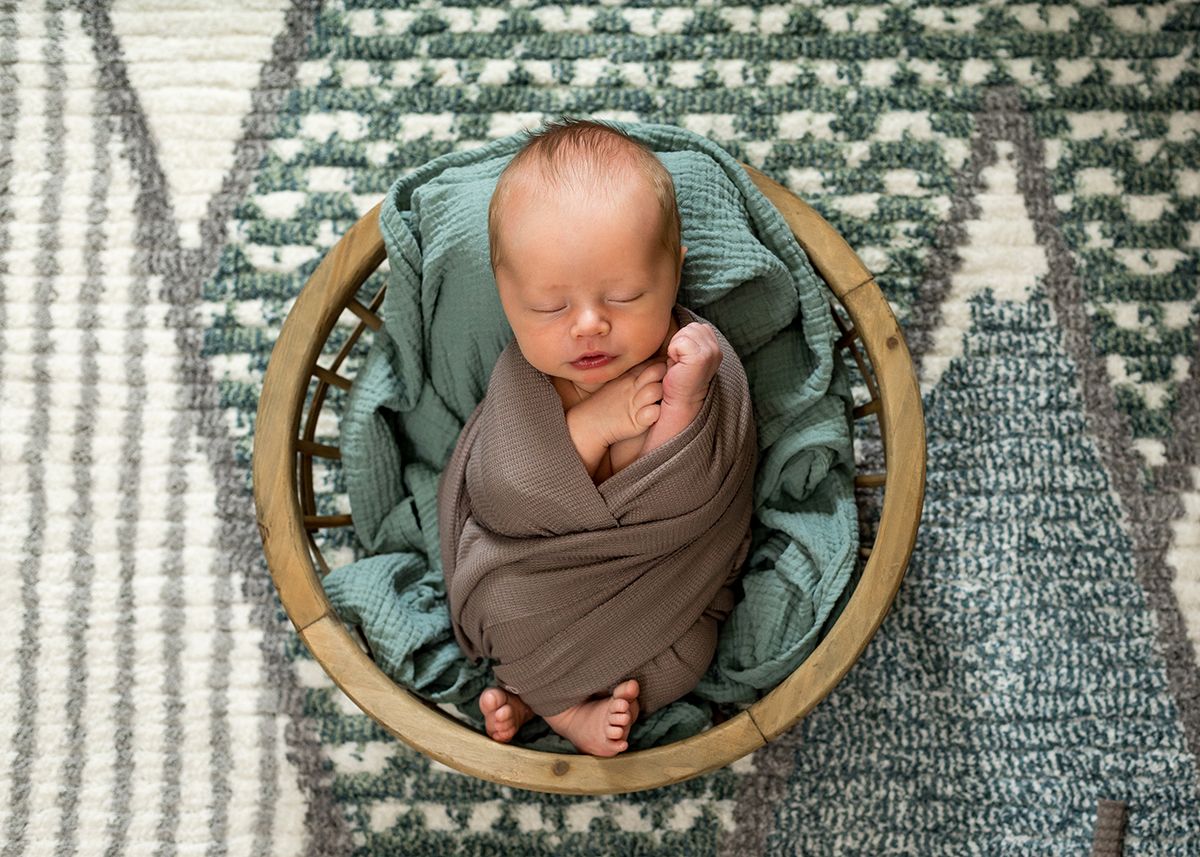 Newborn baby swaddled in gray wrap sleeping peacefully in a round wooden bowl with teal fabric.