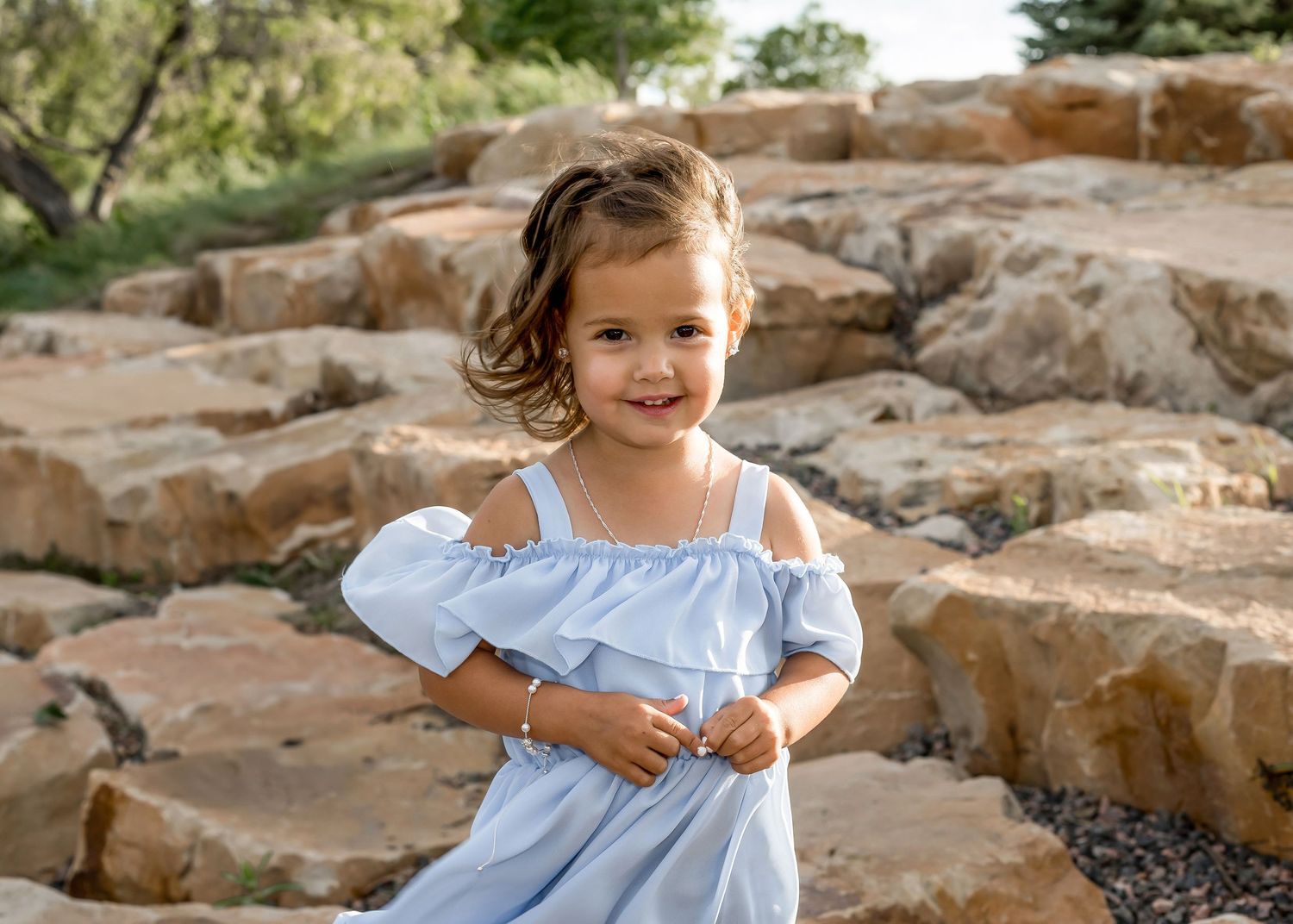 Sweet little girl in a light blue dress posing happily on stone steps outside.