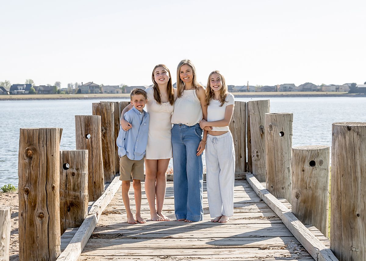 Mother with three children posing on a wooden dock with a serene waterfront backdrop on a sunny day.