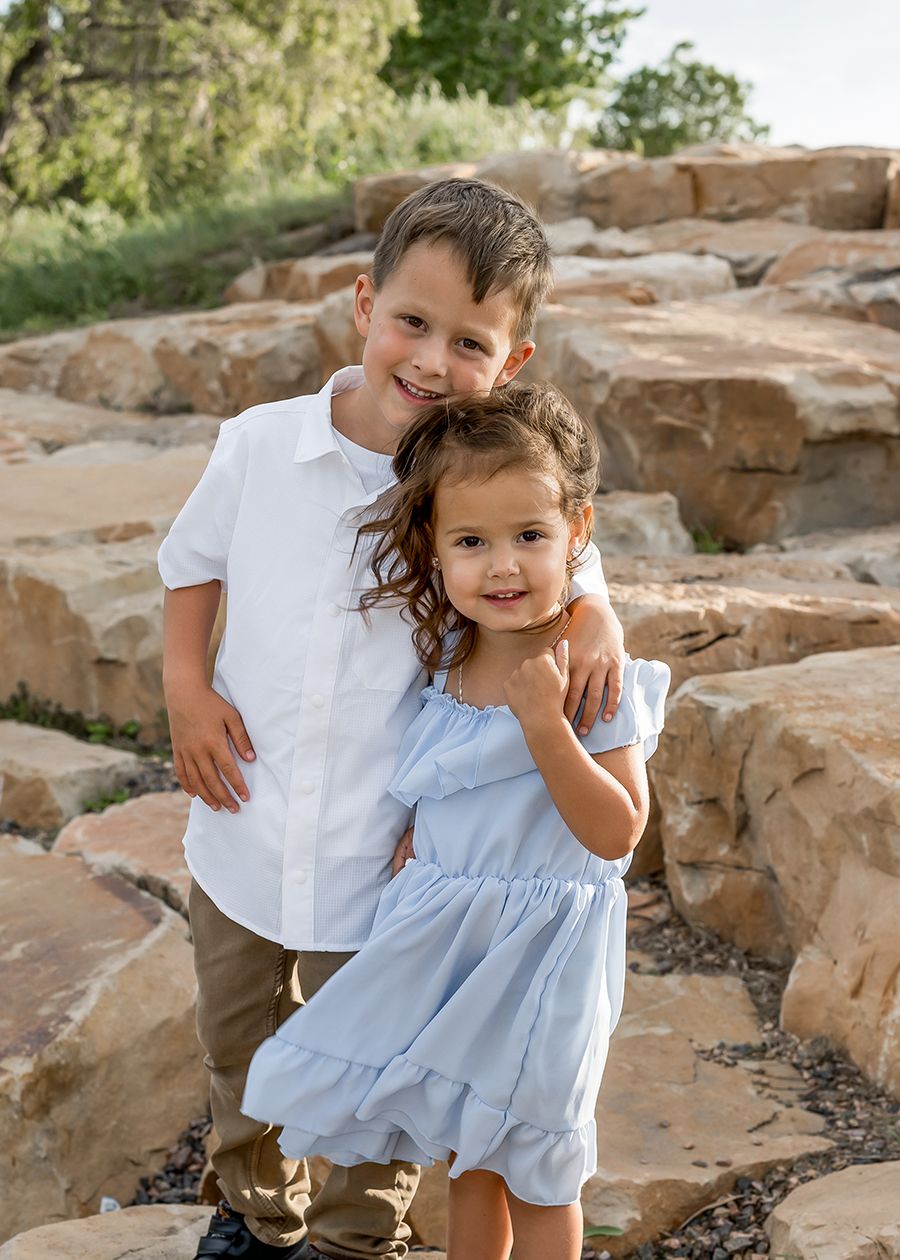 Two young siblings posing on rocky steps outdoors, boy in white shirt hugging girl in light blue dress.
