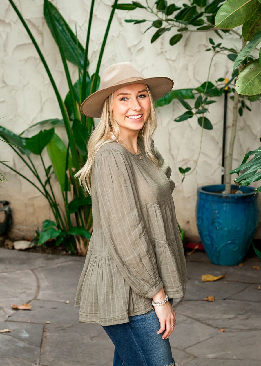 Smiling blonde woman in olive babydoll top and tan wide-brim hat posing outdoors near tropical plants.