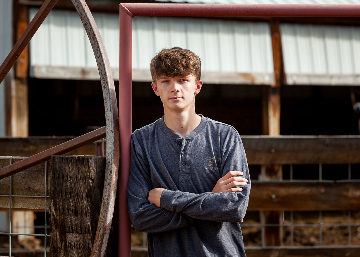 Teen boy in navy henley leans against rustic metal farm gate with crossed arms.