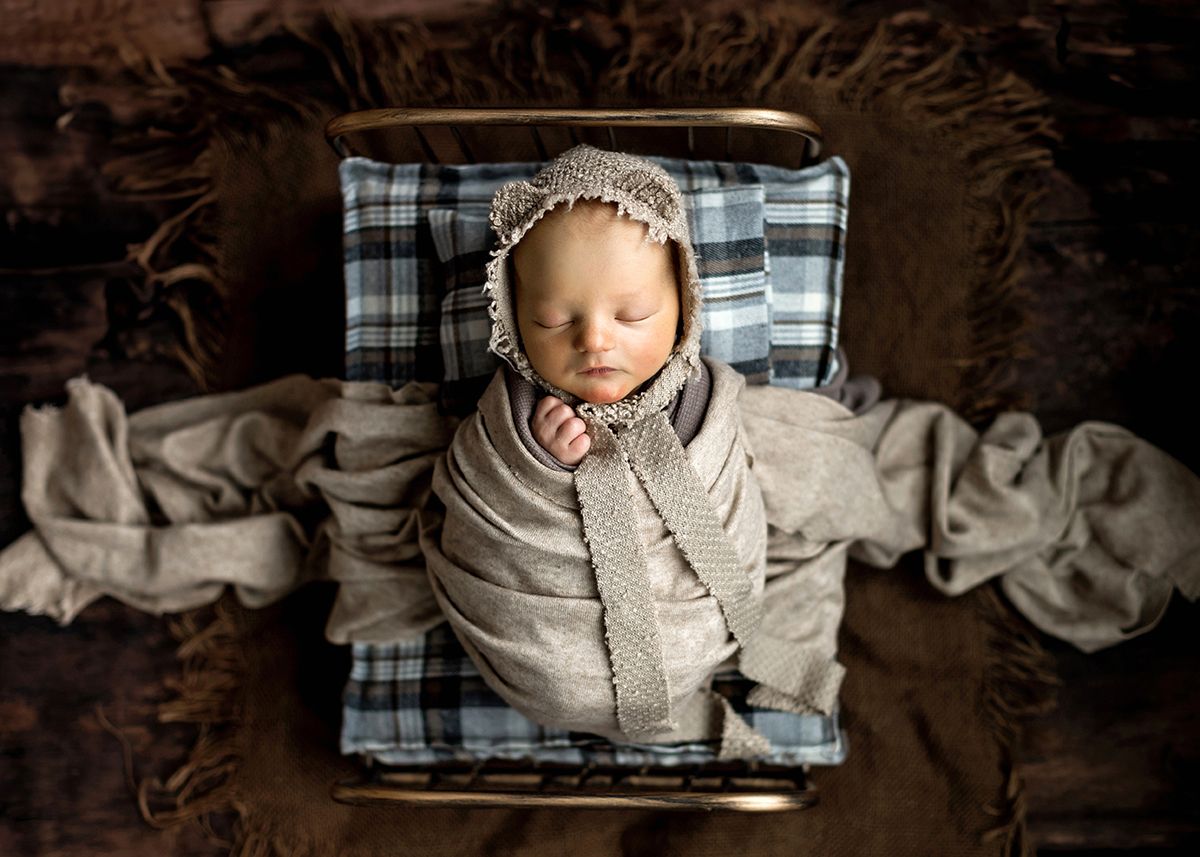 Peaceful newborn baby swaddled in gray knit wrap wearing a bonnet, resting in a vintage metal bed.