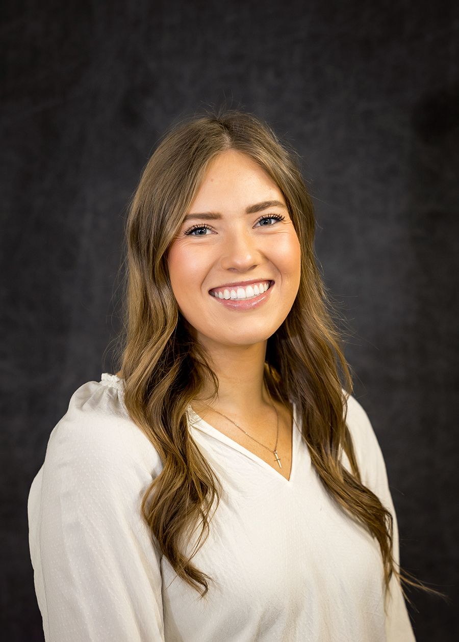 Smiling young woman with long brown hair wearing white blouse, professional headshot on dark background.