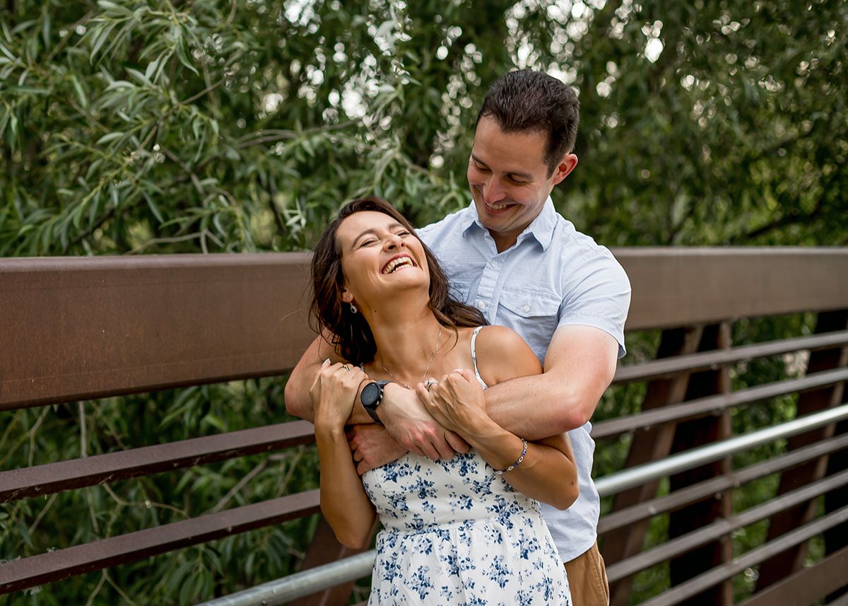 Happy couple embracing on a wooden bridge surrounded by lush green trees, sharing a joyful moment outdoors.