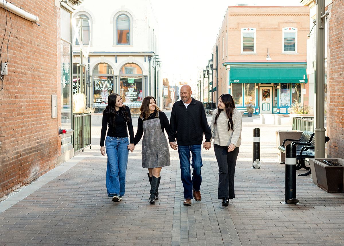 Family of four walks hand-in-hand down a charming brick pedestrian street lined with colorful storefronts.