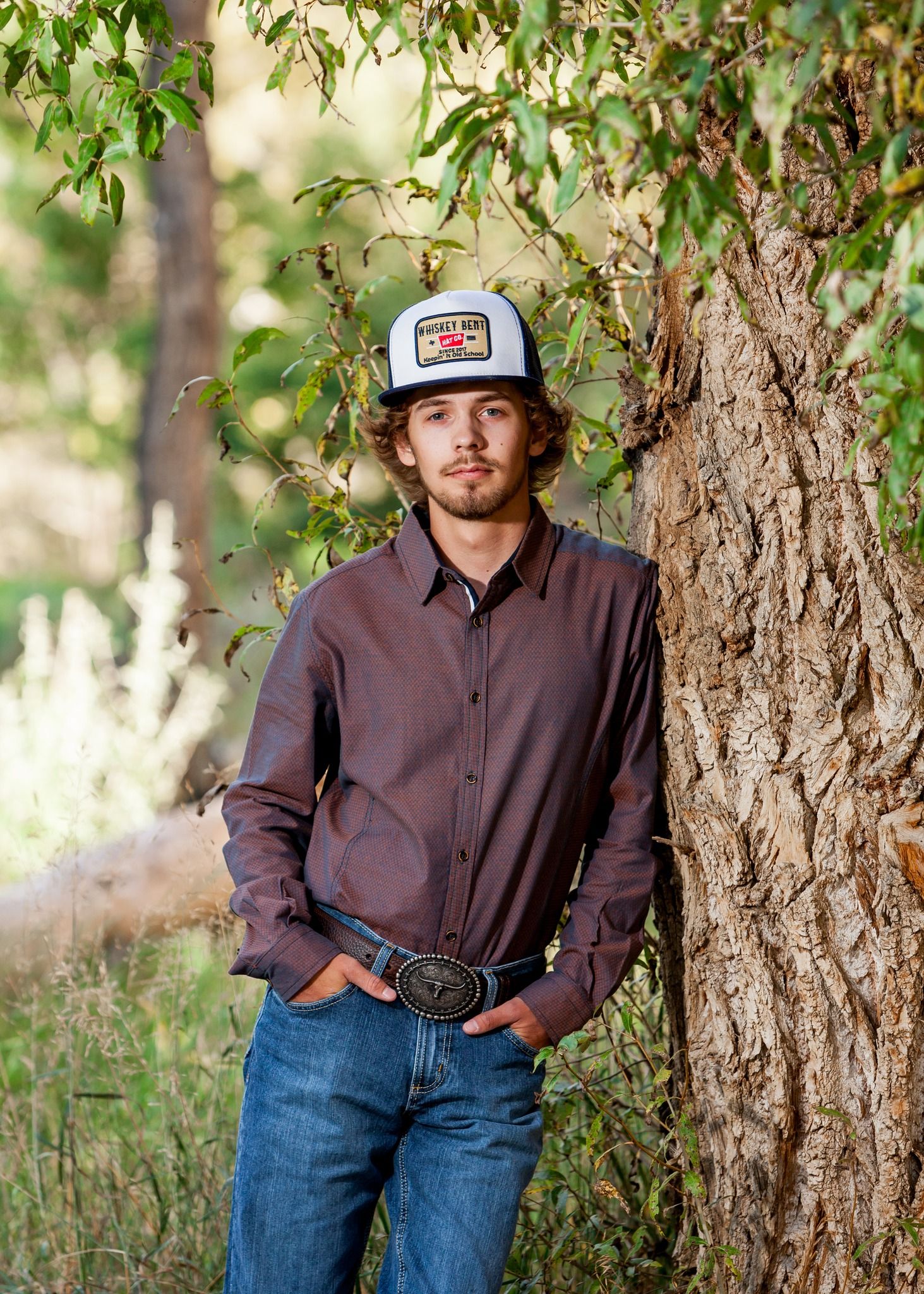 Young man in trucker hat and dark shirt leans against tree trunk outdoors in a rural setting.