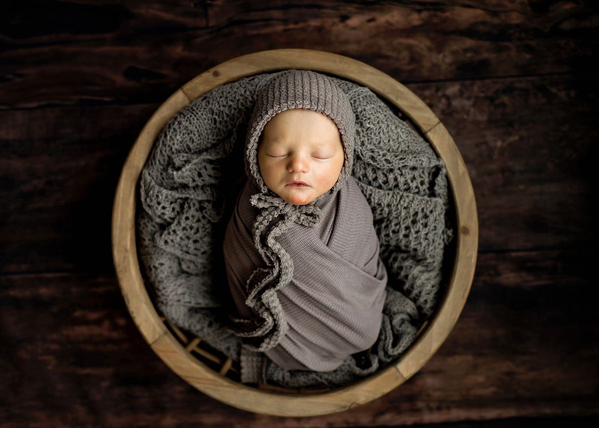 Sleeping newborn baby wrapped in gray swaddle with knit bonnet nestled in a rustic wooden bowl.
