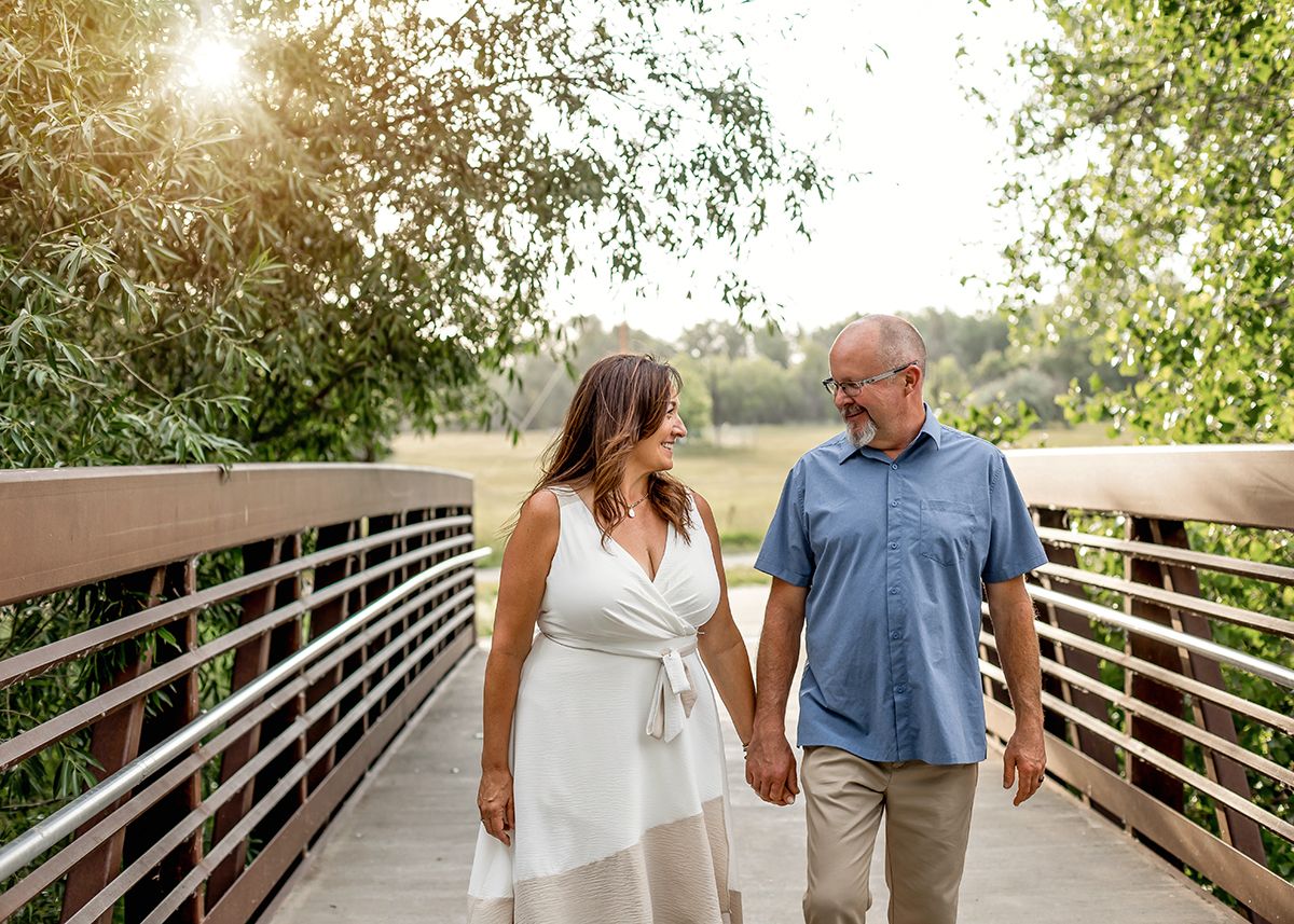Couple holding hands and smiling on a wooden bridge surrounded by lush green trees in warm sunlight.