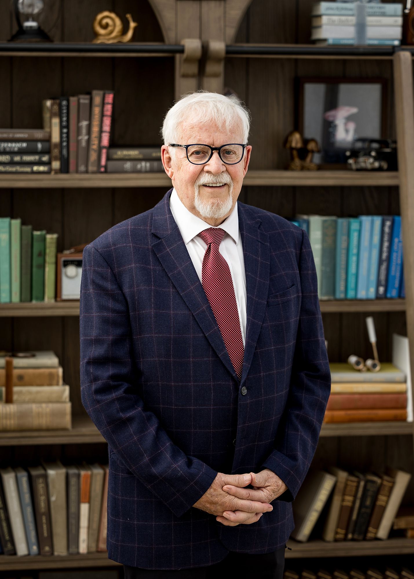 Elderly man with white hair and glasses wearing a navy plaid suit and red tie standing in front of a bookshelf.