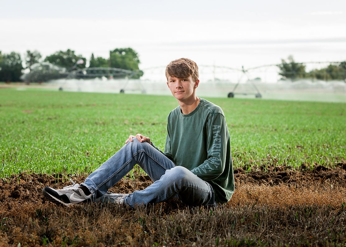Teen boy in green shirt sits relaxed on field edge with irrigation sprinklers behind.