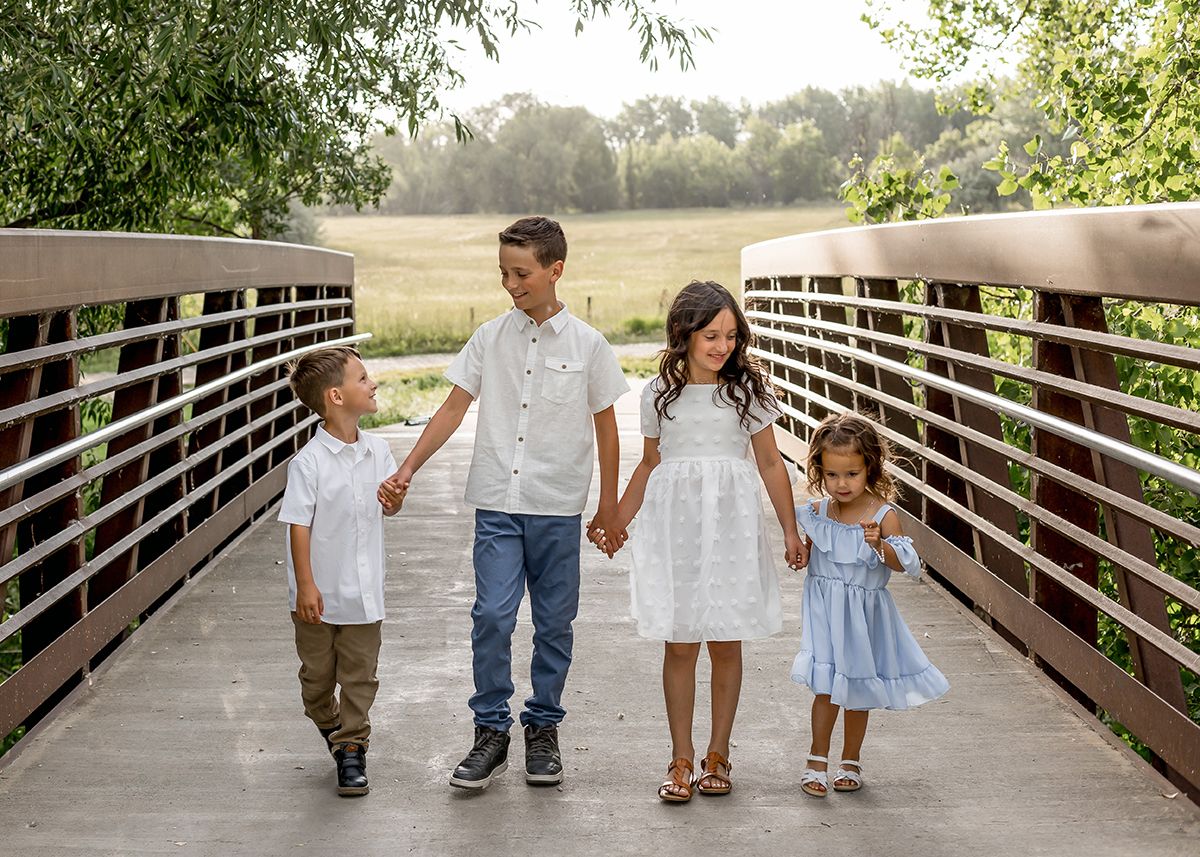 Four children holding hands on a wooden bridge, dressed in white and light blue outfits, surrounded by lush greenery.