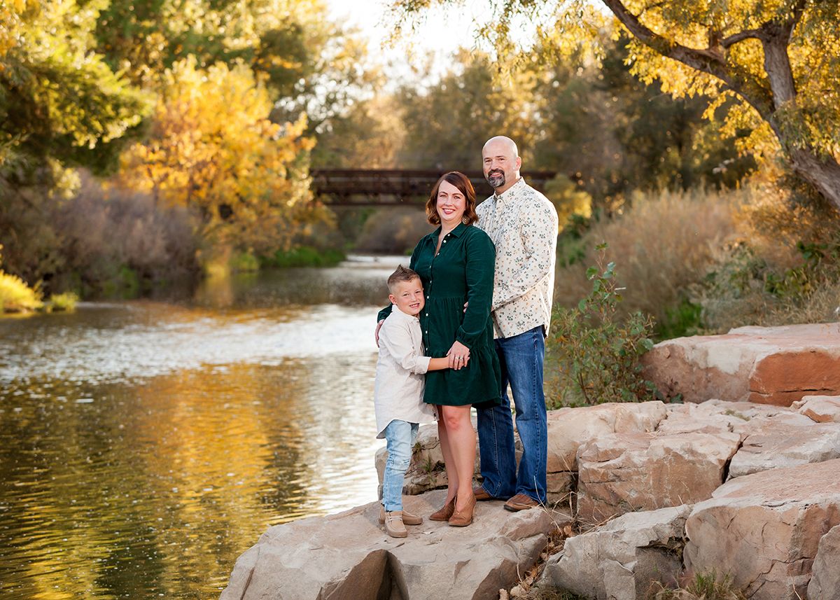 Pregnant family of three posing on rocks beside a golden autumn river.
