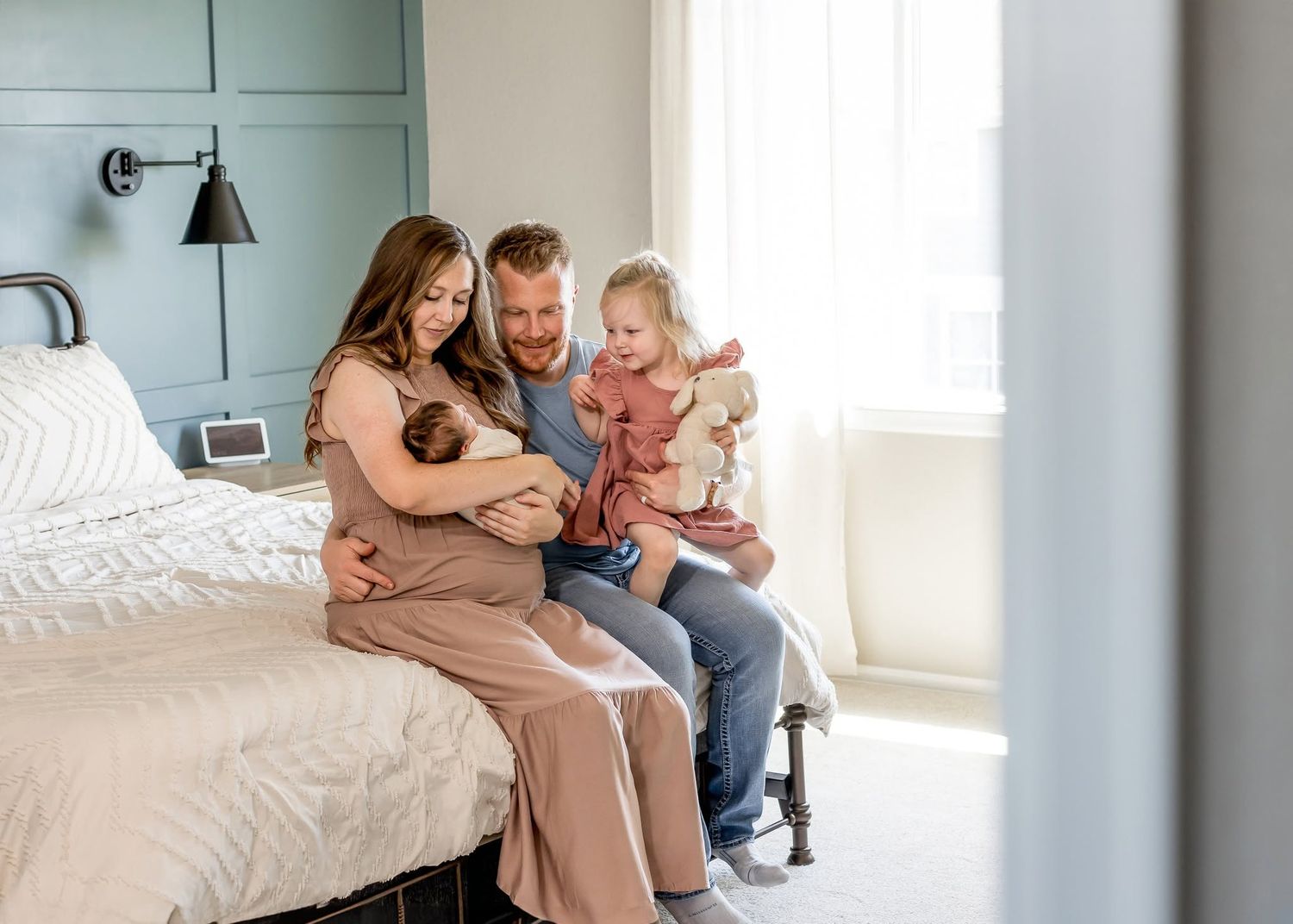 Family of four sitting on bed, parents smiling at newborn baby while young daughter holds stuffed animal.