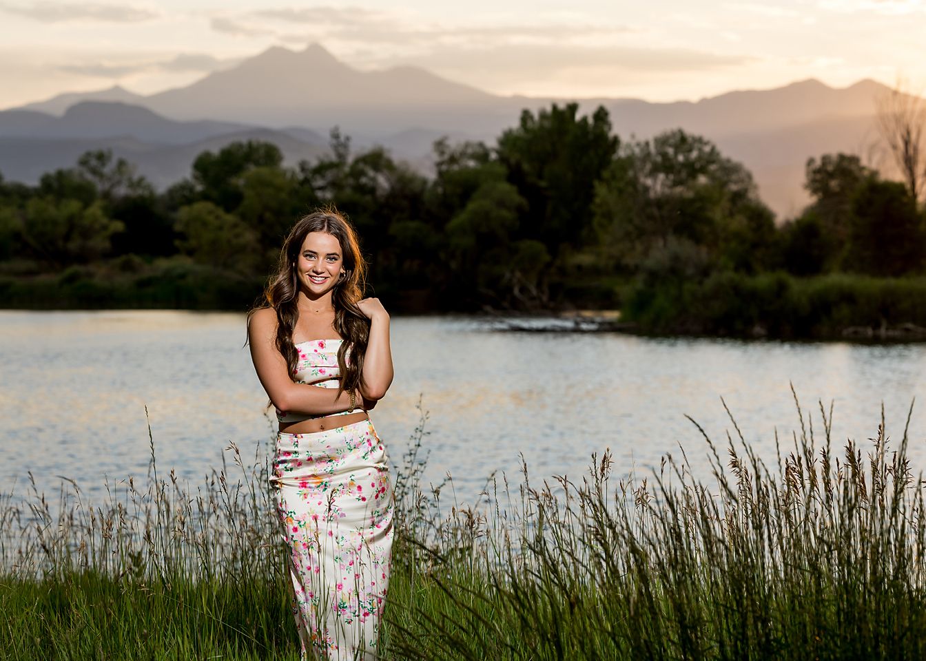 Smiling teenage girl in floral outfit posing near a lake with mountain views at sunset for senior photos.
