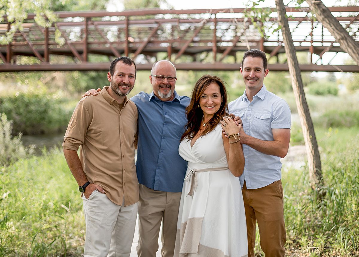 Group of four adults smiling together outdoors with a rustic bridge and greenery in background.