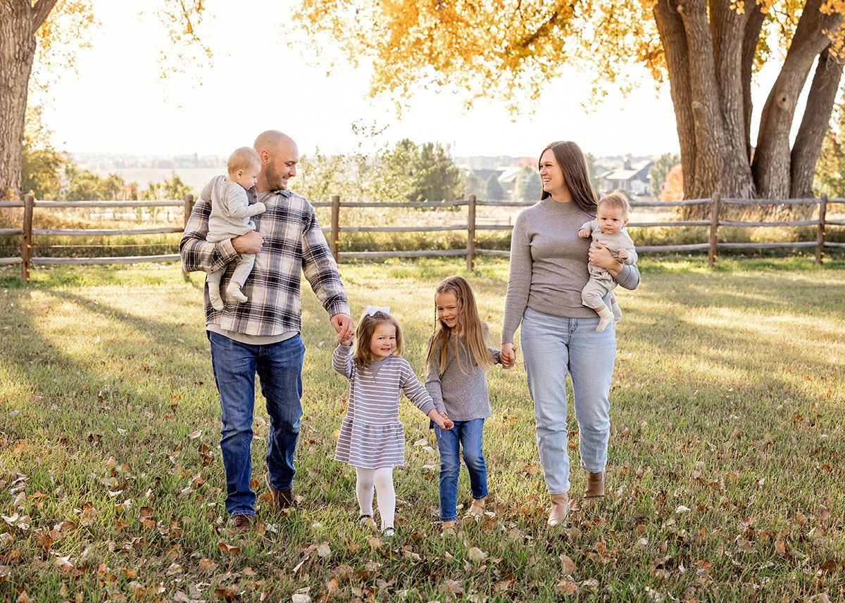 Family of five walking outdoors in autumn park, parents carrying toddlers while two girls hold hands between them.