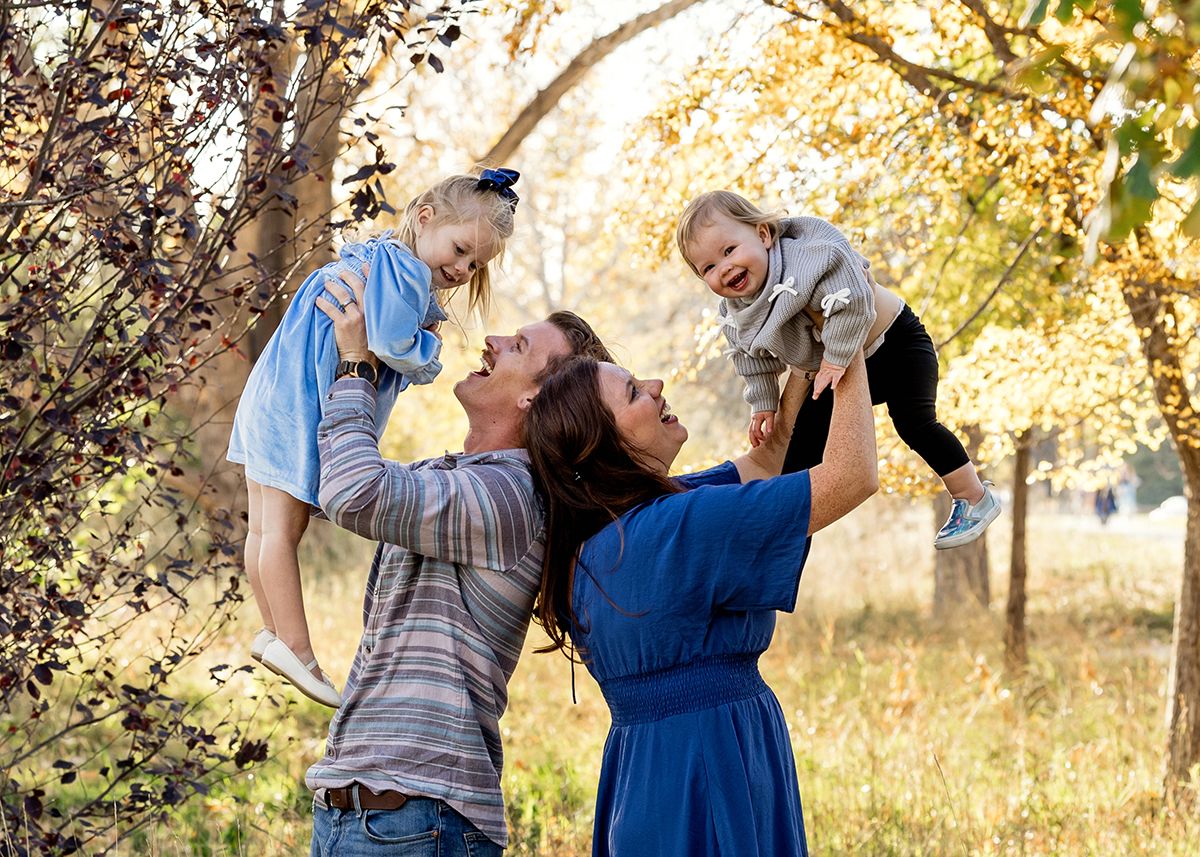 Joyful parents lifting two laughing toddlers among golden autumn trees outdoors.