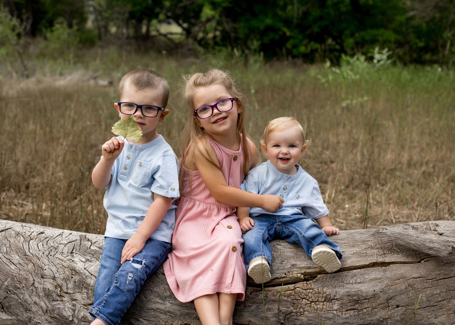 Three young siblings sitting on a log in a field, two wearing glasses, smiling.