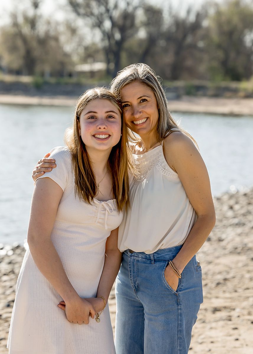 A mother and teenage daughter smile together on a sunny lakeside beach.