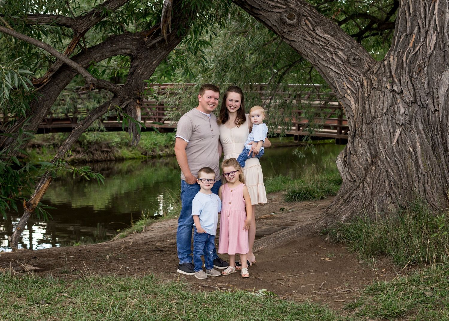 Family of five smiling under a large tree near a peaceful pond and wooden bridge.