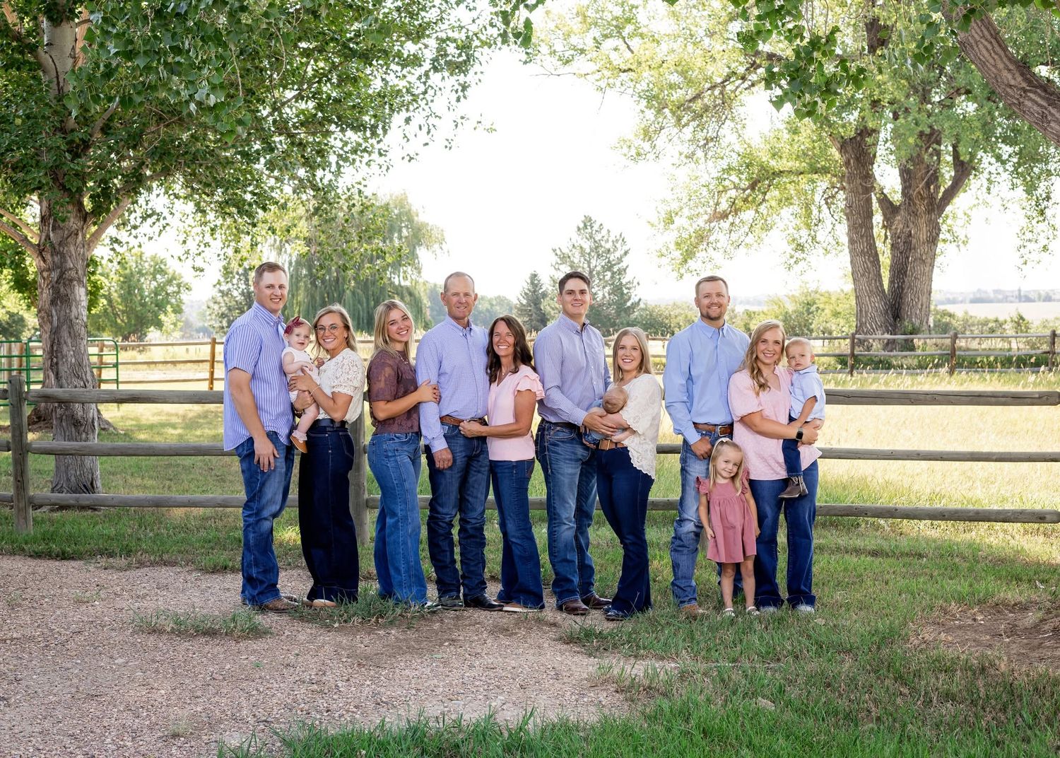 Large family group portrait outdoors on a sunny day, posing together near a wooden fence with green trees.