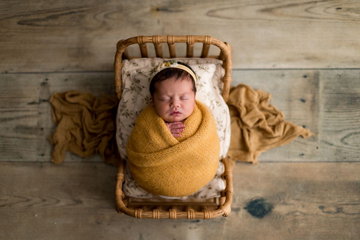 Newborn baby swaddled in mustard yellow wrap sleeping peacefully in a small wicker basket on rustic wood floor.