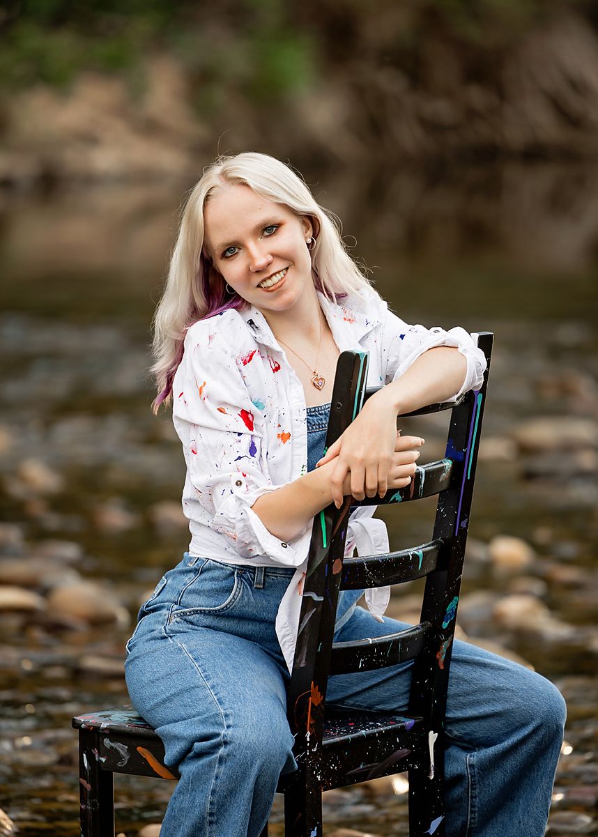 Smiling young woman with blonde hair in paint-splattered shirt and overalls sitting on black chair by rocky creek.