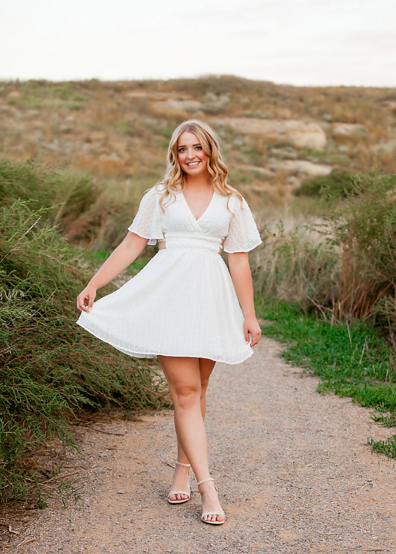 Young woman in white flutter-sleeve mini dress poses on a scenic dirt path surrounded by green shrubs and sandy hills.