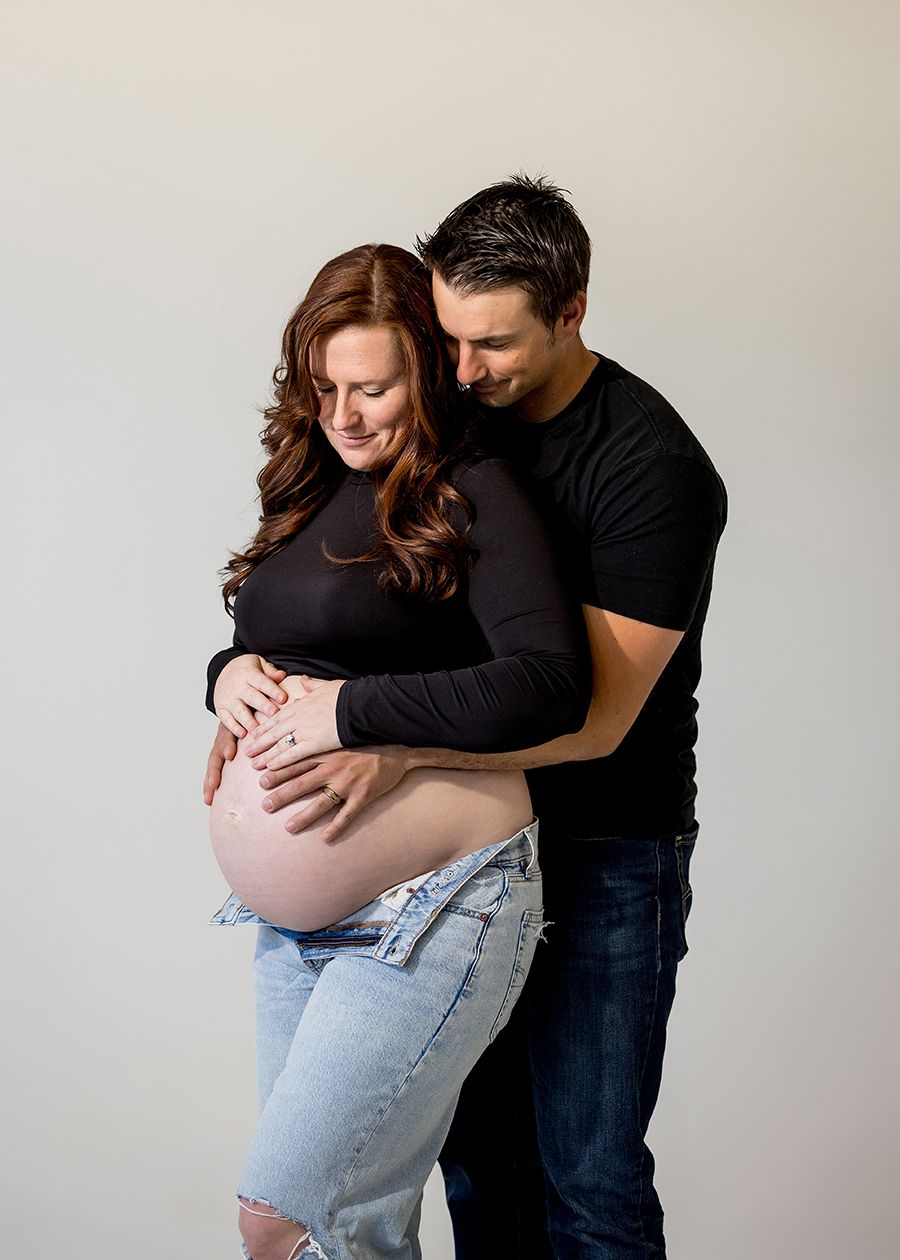 Couple in black tops cradles pregnant belly during maternity photo session against neutral studio background.