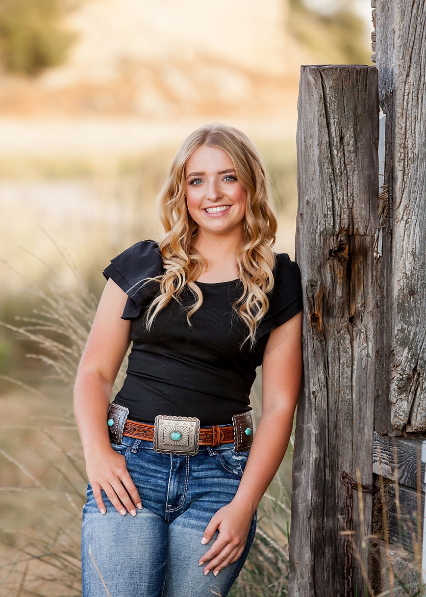 Blonde woman in black top, jeans, and turquoise belt poses by a wooden fence post in a golden field.