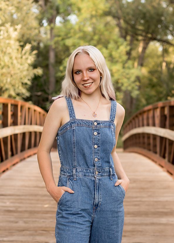 Blonde teen in denim overalls smiles with hands in pockets on a wooden bridge in a forest park.