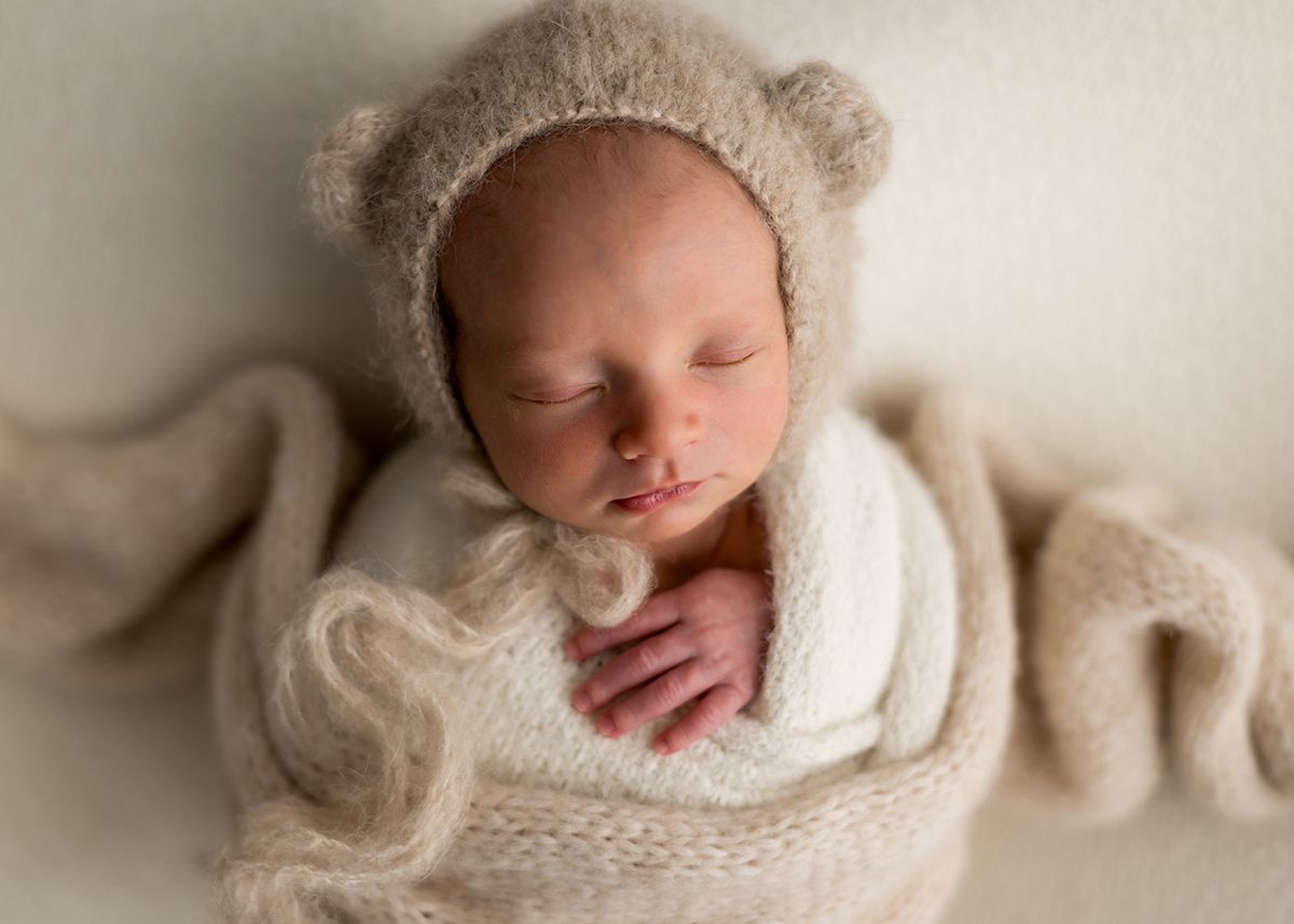 Sleeping newborn baby wrapped in white knit swaddle wearing a beige bear ear bonnet.