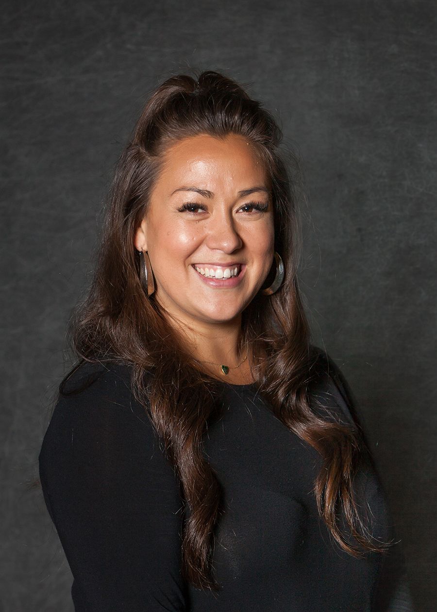 Smiling woman with long brown hair wearing a black top, professional headshot on dark gray background.