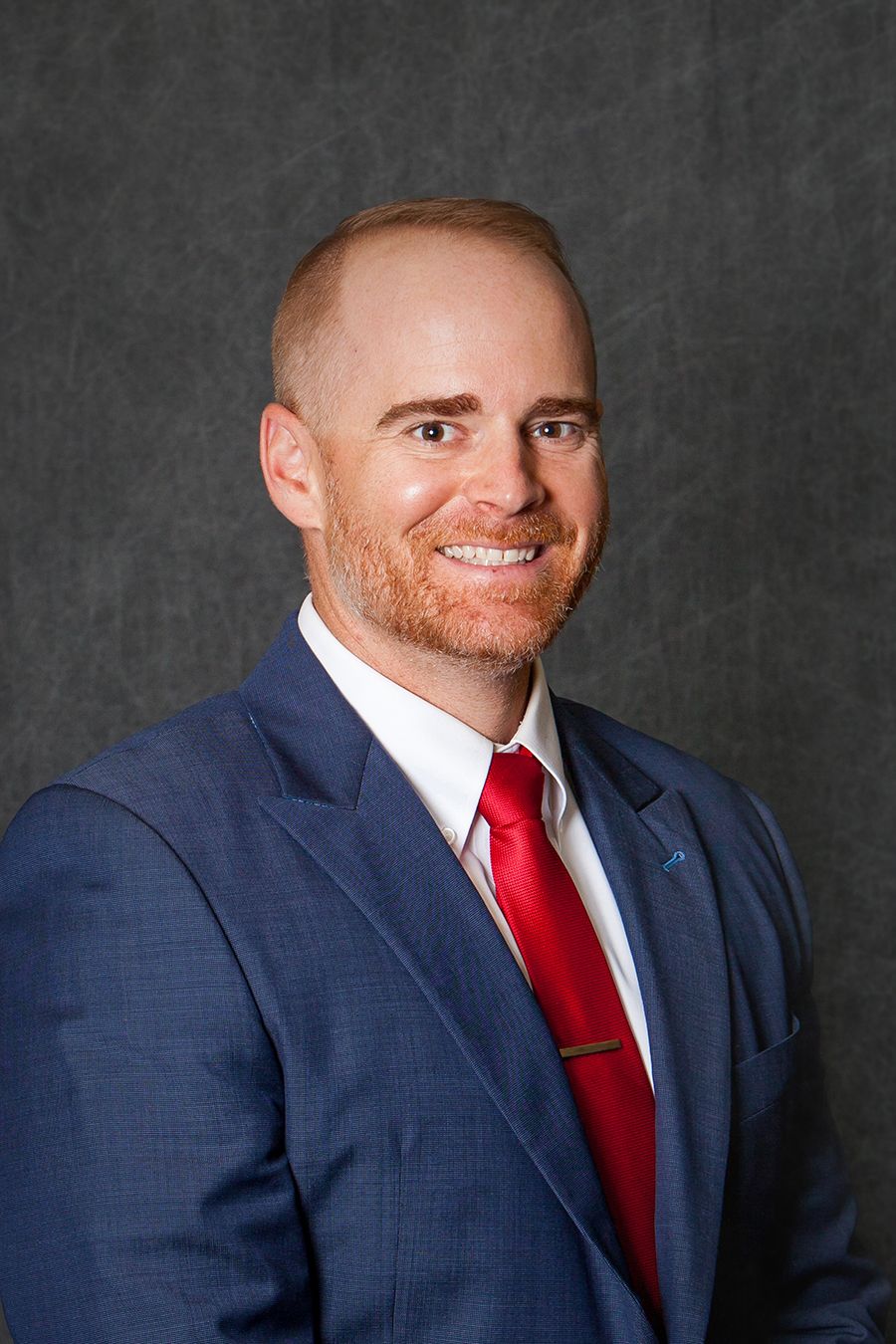 Professional man in blue suit and red tie with short reddish hair smiling against a gray background.