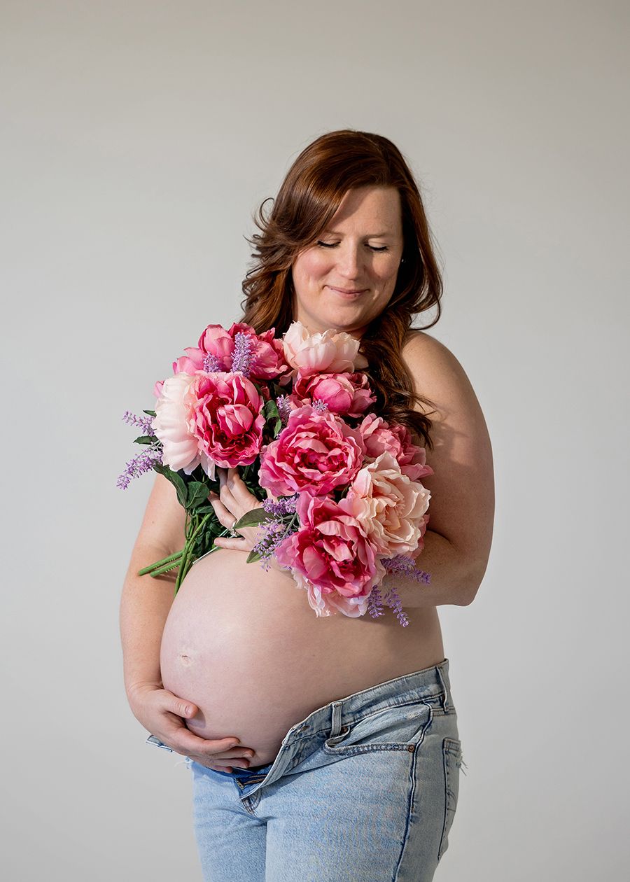 Pregnant woman holding pink peonies bouquet, wearing jeans, cradling bare baby bump in a studio maternity photo.