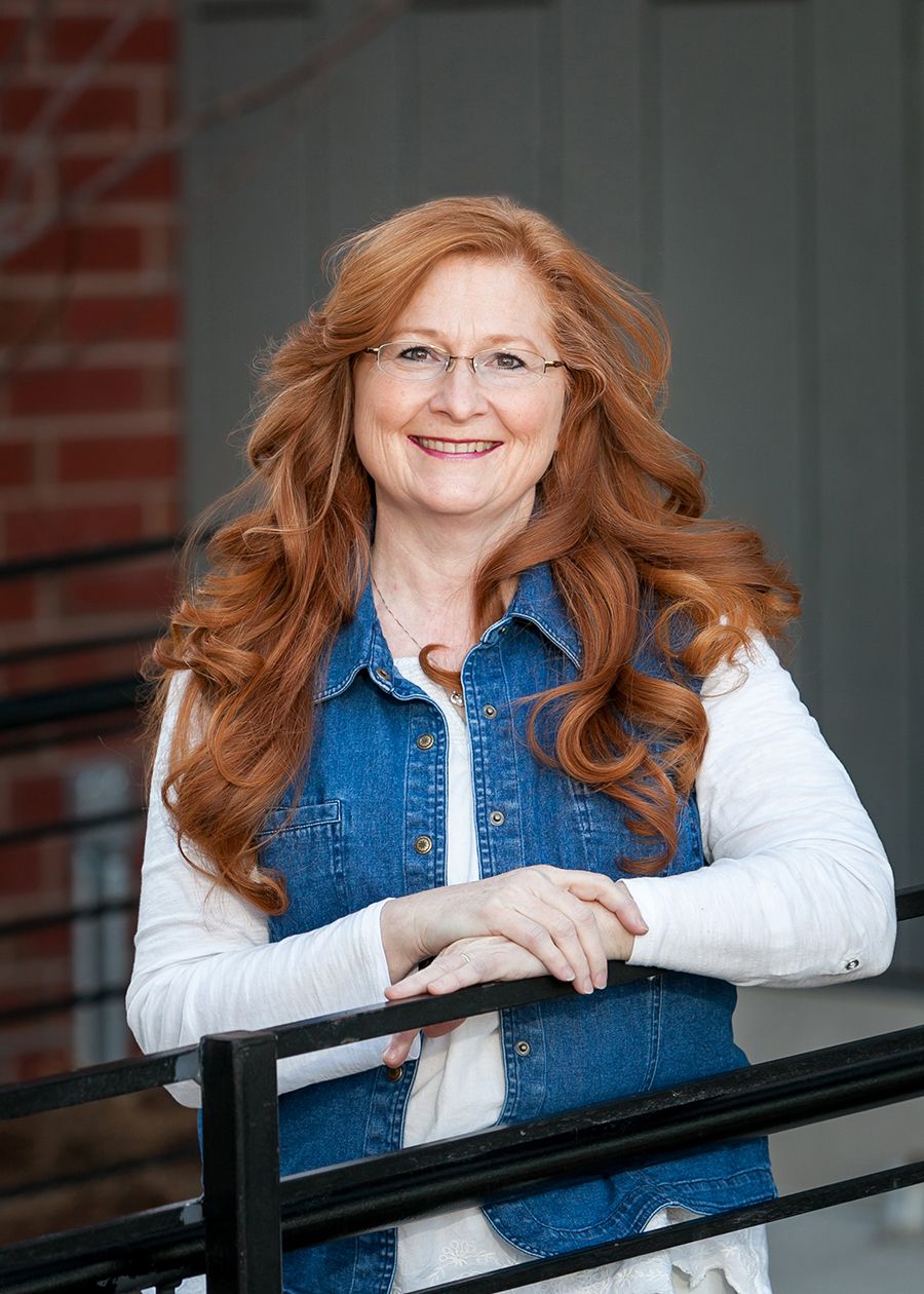 Smiling woman with long red hair and glasses wearing a denim vest over white shirt, leaning on a black railing.