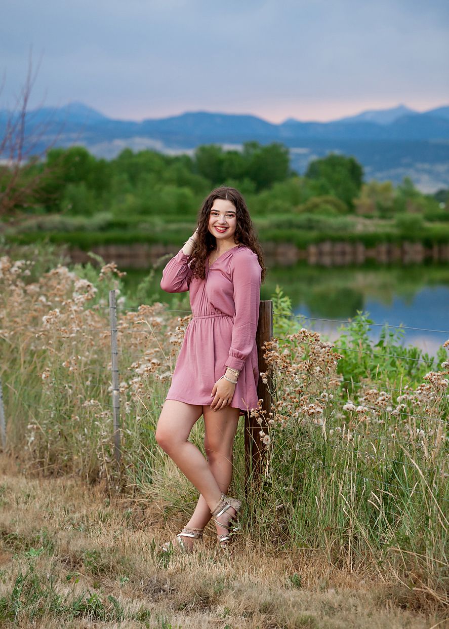 Young woman in pink dress poses near lake with mountain backdrop for outdoor senior photo session.