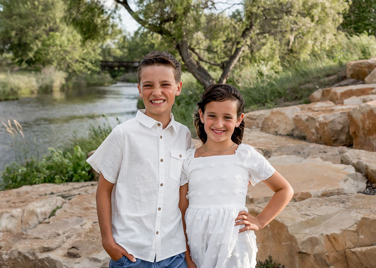 Two smiling children in white outfits pose on rocky terrain near a river surrounded by lush green trees.