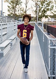 Teen boy in football jersey holds ball while walking on stadium bleacher ramp.