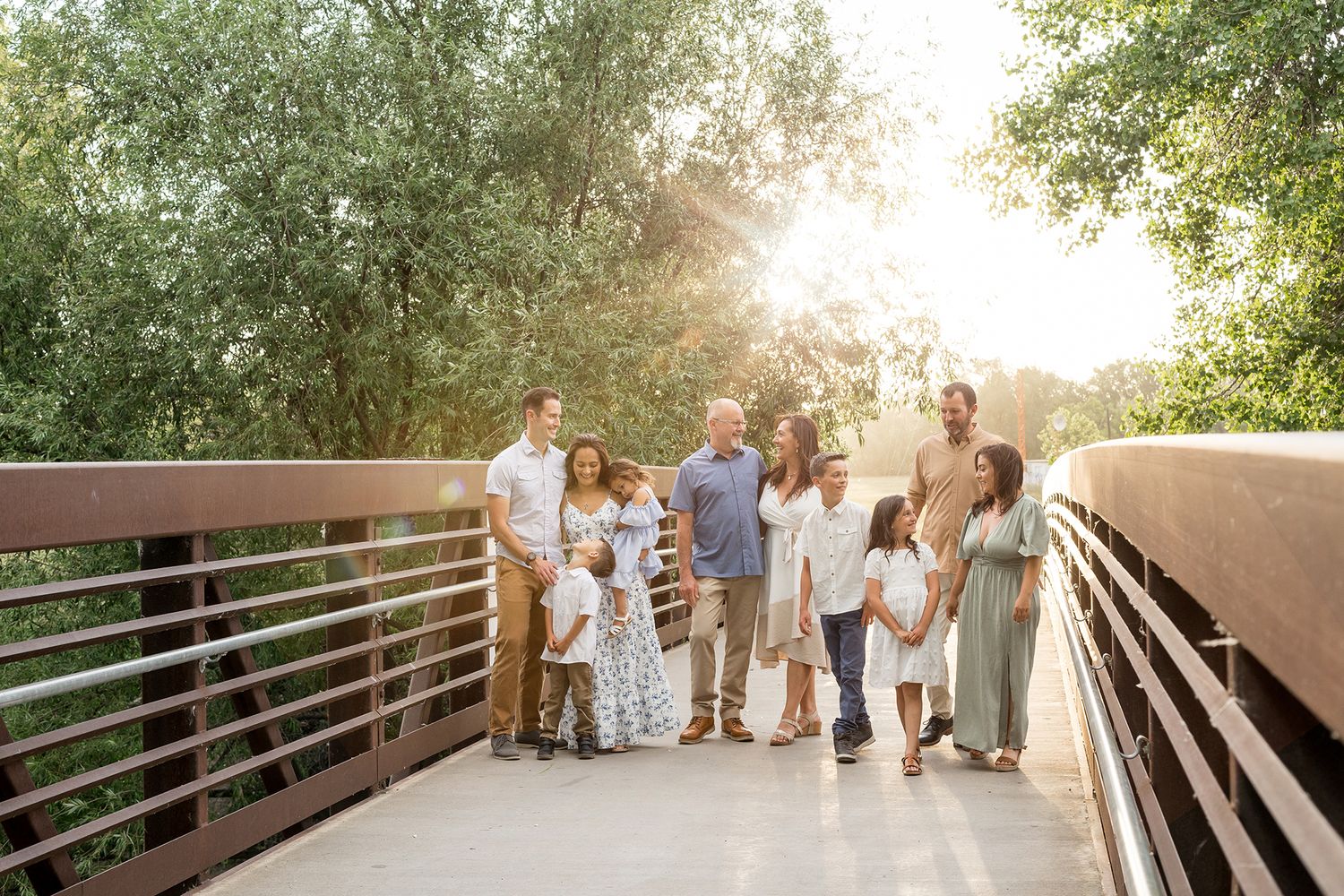 Large family posing together on a bridge surrounded by lush green trees with golden sunlight in the background.