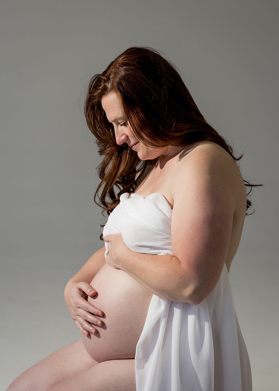 Pregnant woman in white strapless dress cradling her baby bump, looking down tenderly in a studio portrait.