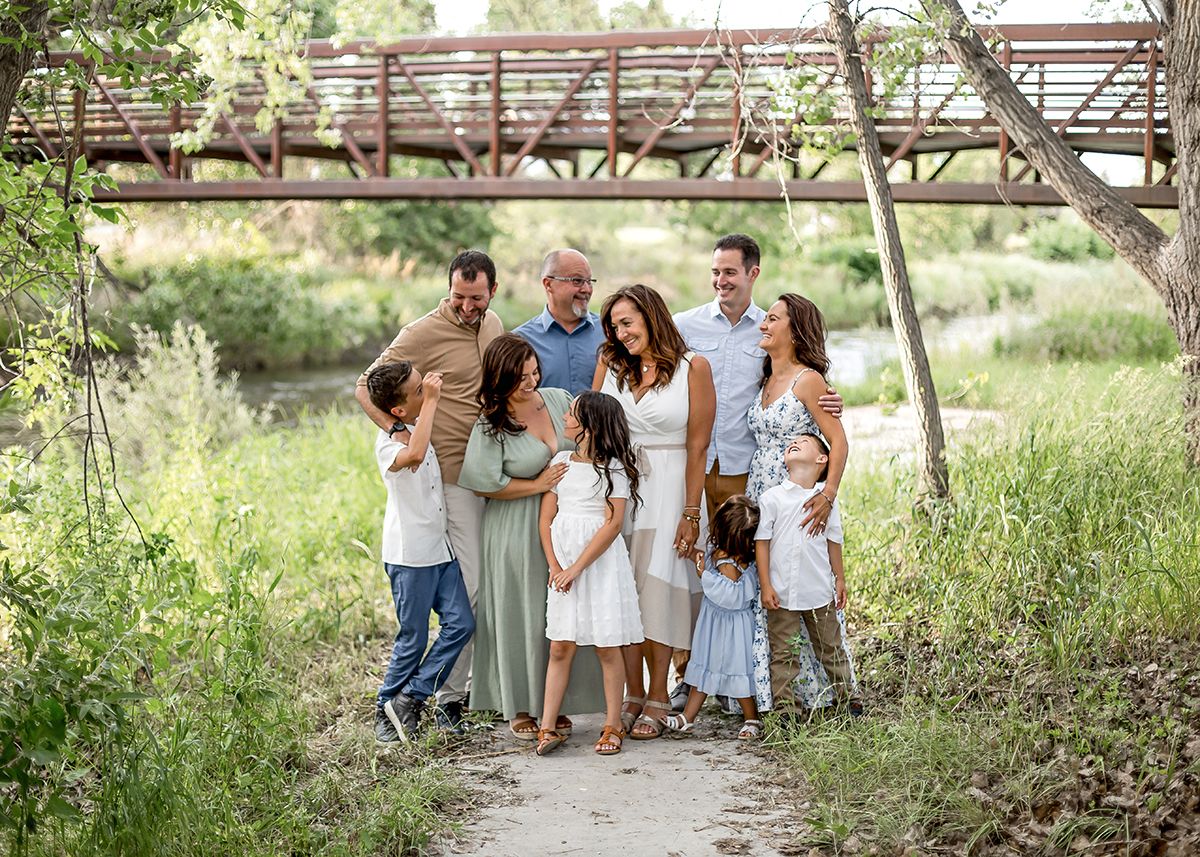 A large multigenerational family poses together on a scenic outdoor path surrounded by lush greenery near a bridge.