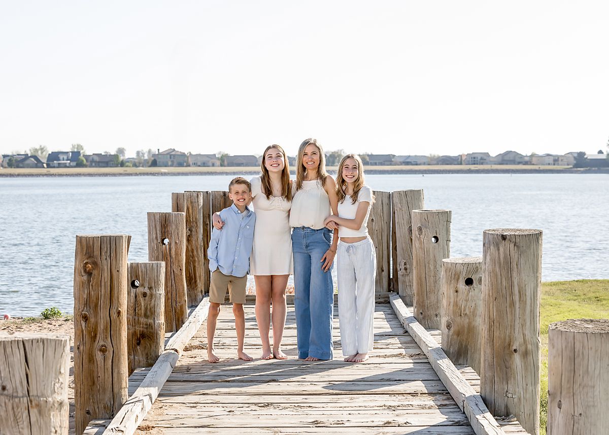 Four siblings pose together on a wooden dock by the water on a sunny day.