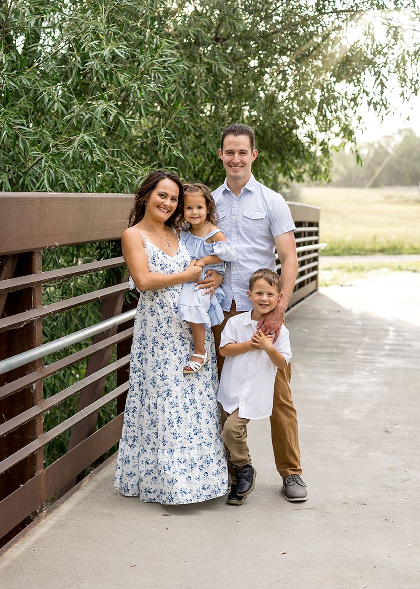 Family of four posing on a wooden bridge surrounded by lush green trees in summer.