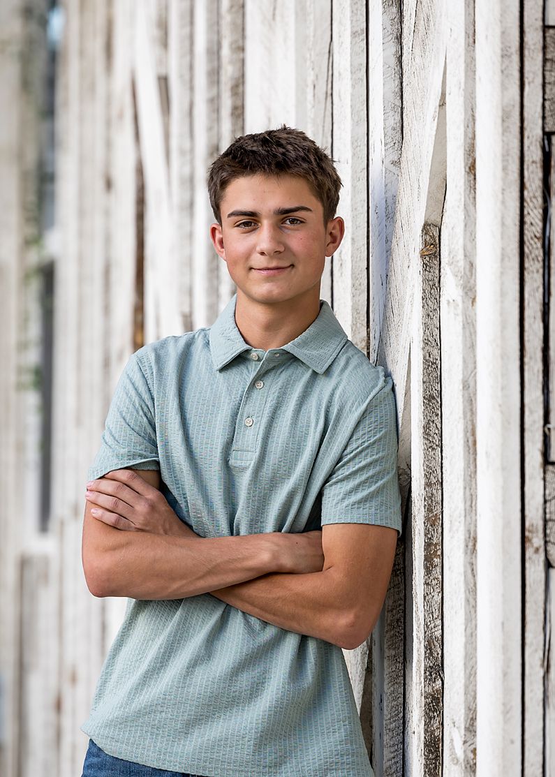Teenage boy in light blue polo shirt leaning against a rustic white wooden wall with arms crossed for senior portrait.