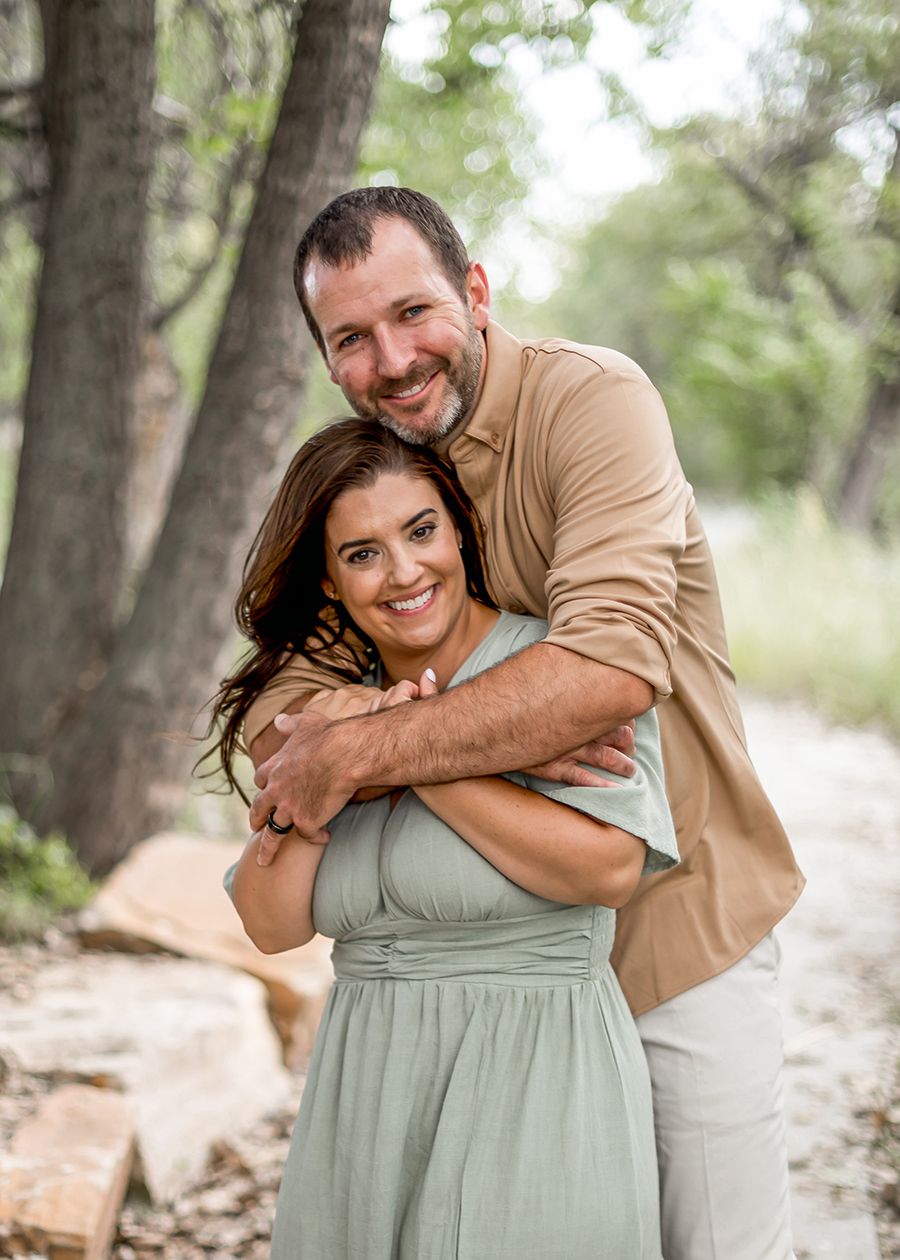 Man in tan shirt embracing smiling woman in sage green dress outdoors among trees.