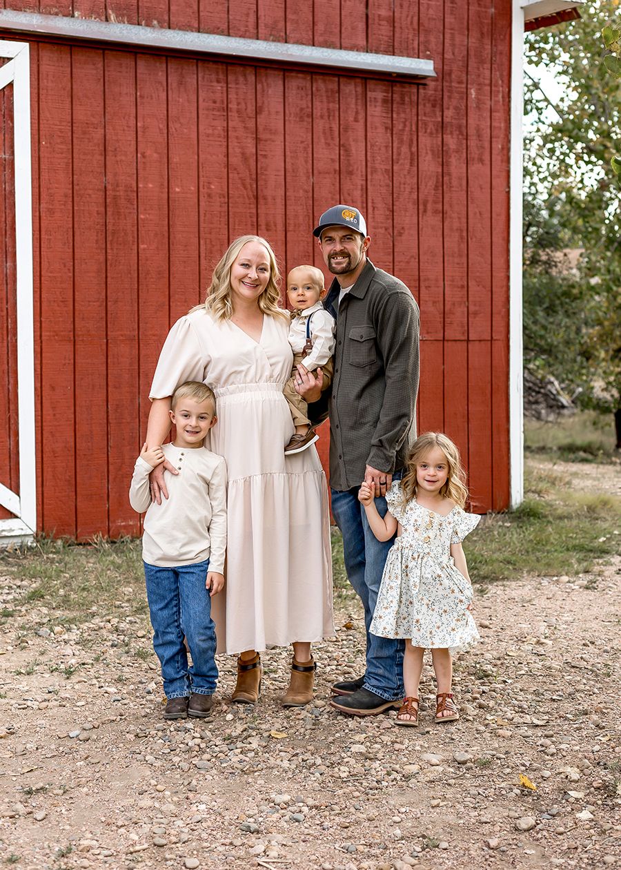 Happy family of five posing together in front of a classic red barn on a farm.